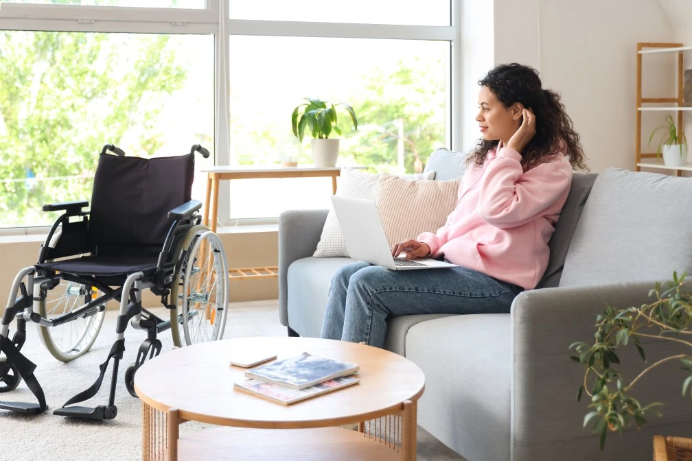 Disabled woman sitting in living room on sofa next to wheelchair using laptop to contact H3 Care