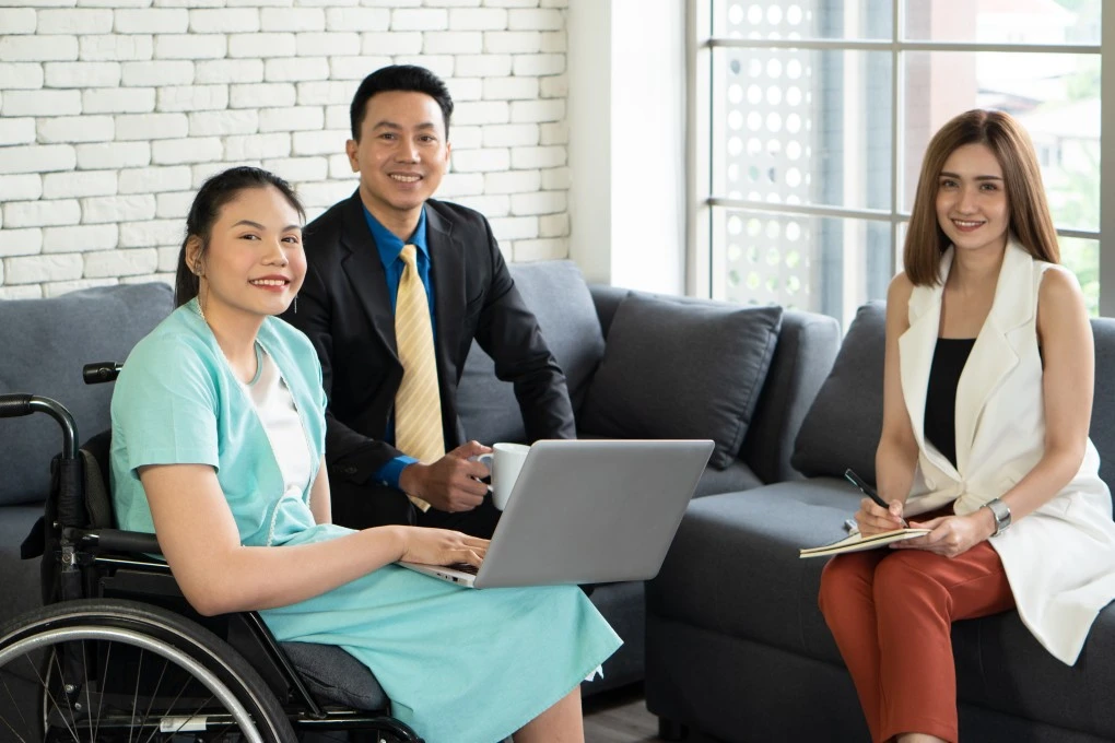 Disabled woman in wheelchair holding laptop and smiling with two support workers